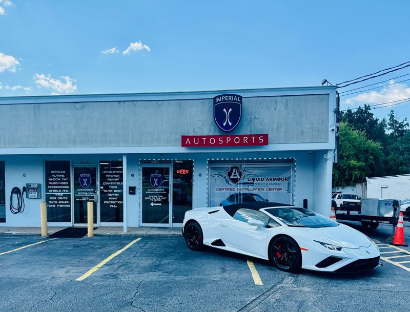 White convertible sports car parked in front of Imperial Autosports shop with blue logo and red 'AUTOSPORTS' sign under a clear blue sky.
