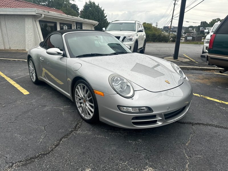 Silver Porsche 911 convertible parked in a parking lot, soft top up, front angled view showing round headlights and logo