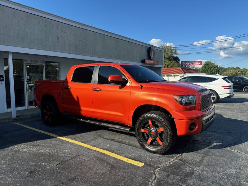 Orange pickup truck parked in a lot in front of a shop, with shadows on the pavement and blue sky above.