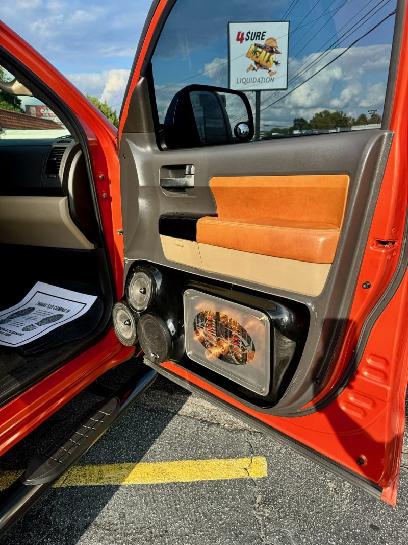 Open orange pickup door showing tan leather interior and a large chrome speaker box with multiple drivers at the bottom of the door panel, on a sunny day by a dealership lot.