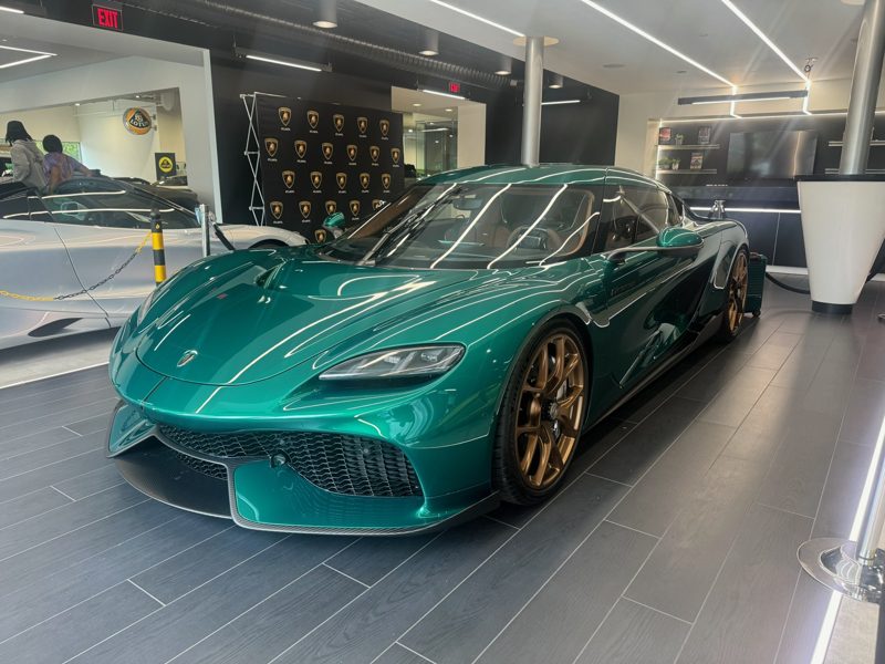 Teal-green exotic sports car on a showroom floor, front angled view with bronze wheels and glossy finish.