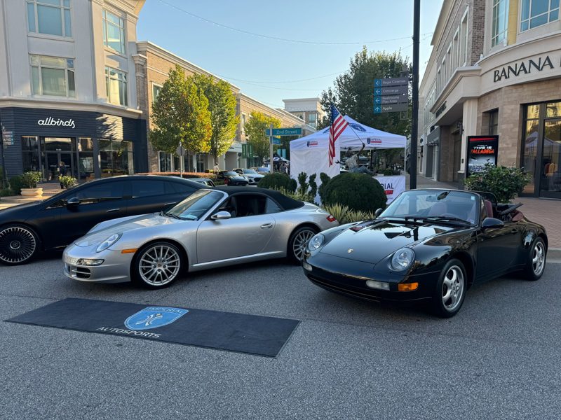 Two Porsche convertibles parked on a pedestrian street during a car show, a silver coupe on the left and a black convertible on the right, with storefronts and a tent in the background.