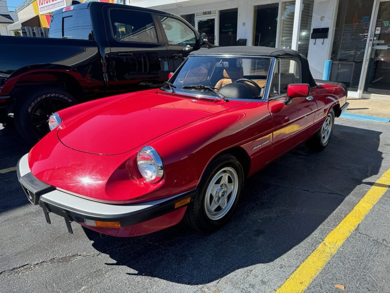 Red vintage convertible with black soft top parked in a lot, tan interior visible through the windshield, chrome bumper accents.