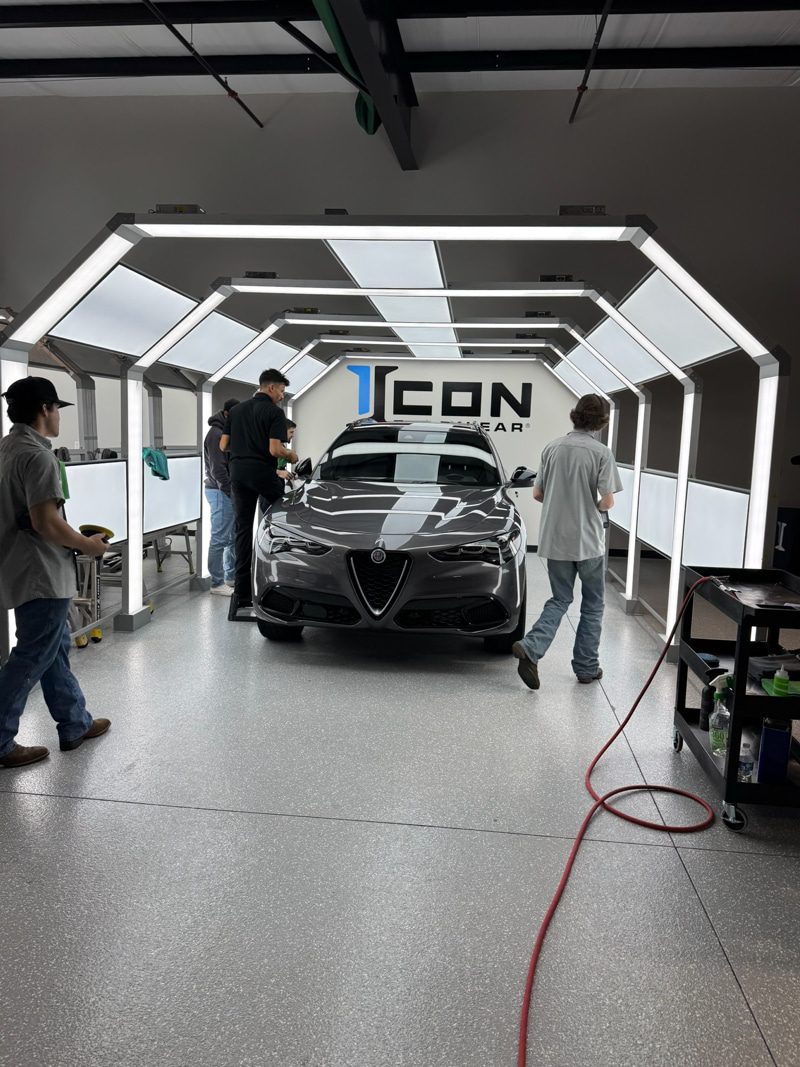 Grey luxury sedan centered in a bright, angular-lit detailing bay with technicians nearby.