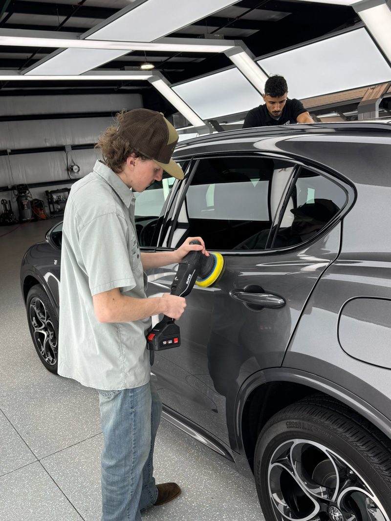 Young man polishing a gray SUV with a handheld buffer inside a bright workshop, another person on the roof in the background.