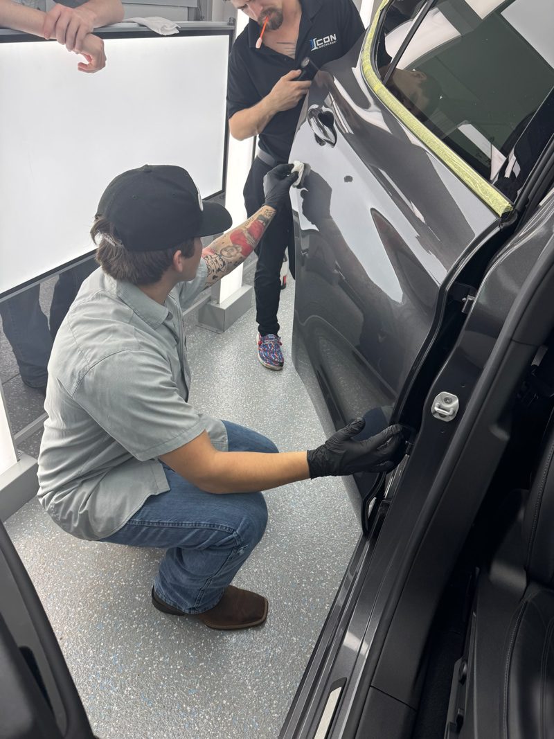 Two technicians work on the side of a dark gray SUV in a workshop; one kneels and uses a tool near the door while the other stands nearby holding a panel or tool. Masking tape lines the window edge.
