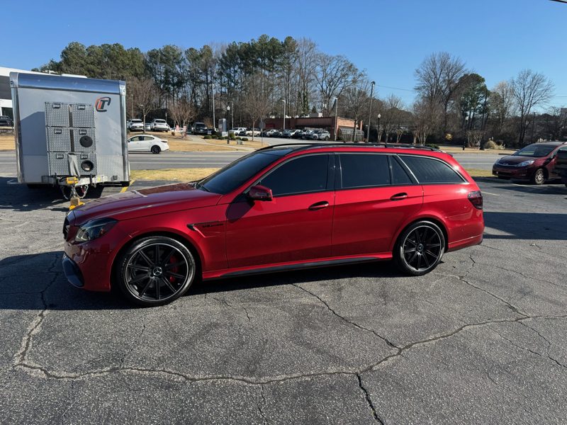 Red Mercedes-Benz station wagon with black alloy wheels parked in a lot, trailer on the left and trees in the background.