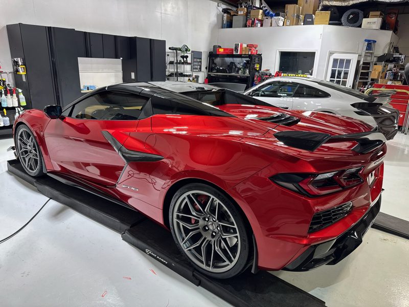 Red mid‑engine Corvette on a service lift in a workshop, black roof, and prominent rear spoiler.