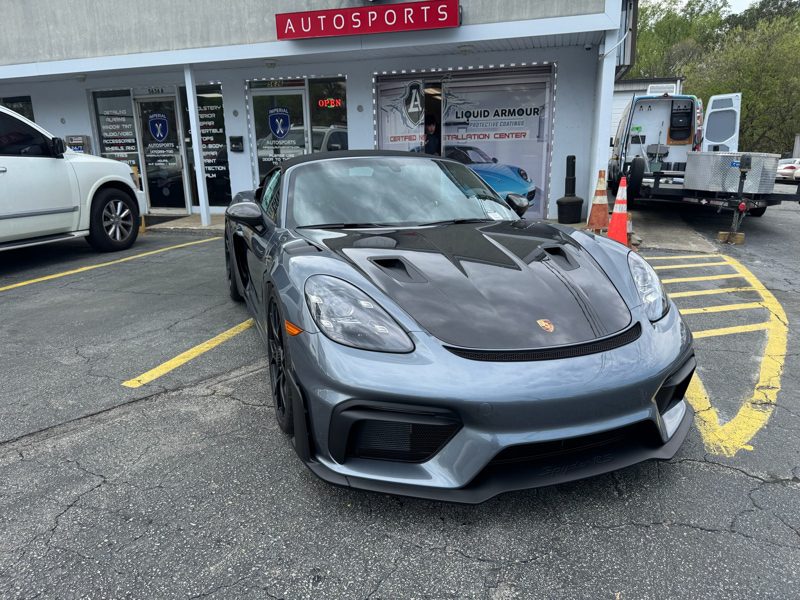 Gray Porsche sports car parked diagonally in front of an Autosports storefront, with a white SUV nearby and a traffic cone visible by the entrance.