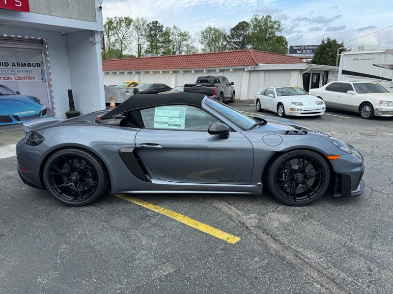 Gray Porsche 718 Spyder RS with black wheels, convertible top up, parked in a lot beside a storefront with a window sticker visible on the passenger window.