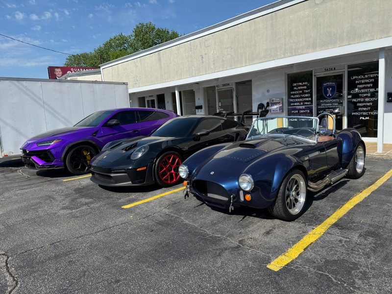 Three sports cars parked in front of a white storefront: a purple coupe on the left, a black car with red wheels in the middle, and a navy blue vintage open‑top roadster on the right.