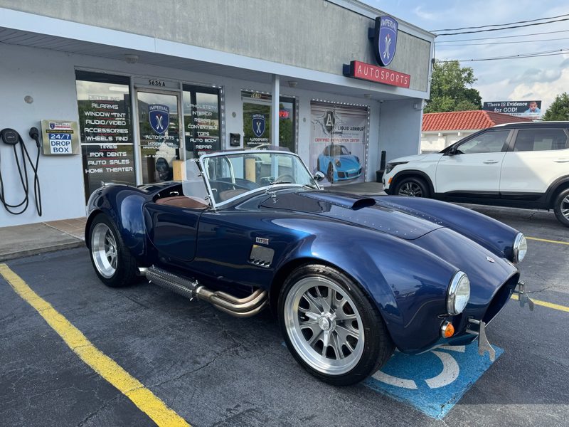 Blue classic convertible sports car with chrome wheels parked outside an auto shop storefront (AUTOSPORTS). Named storefront signage visible in background.