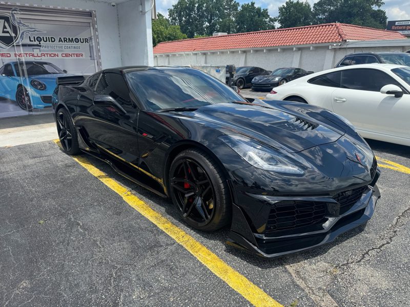 Black Chevrolet Corvette sports car parked in a lot beside a shop, featuring a low stance, glossy finish, and red brake calipers.