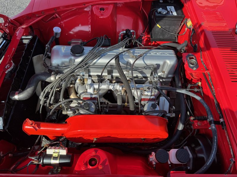 Red car engine bay with a silver inline-four engine, red intake cover, and various hoses and wires visible under the hood.