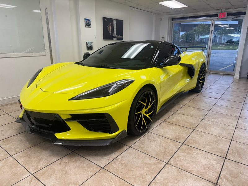 Yellow Chevrolet Corvette C8 in a showroom with a black roof and large black-spoked wheels, front splitter visible.