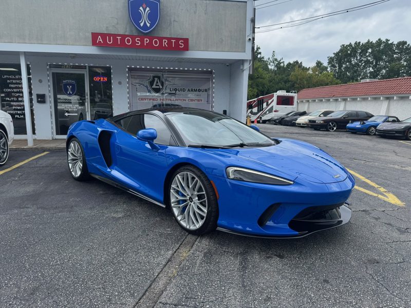 Blue McLaren sports car parked in front of an autosports shop with a gray facade and open doors signage visible.