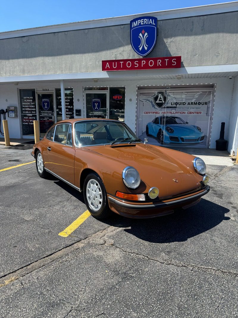 Brown classic Porsche 911 parked in front of Imperial Autosports shop on a sunny day in the lot.