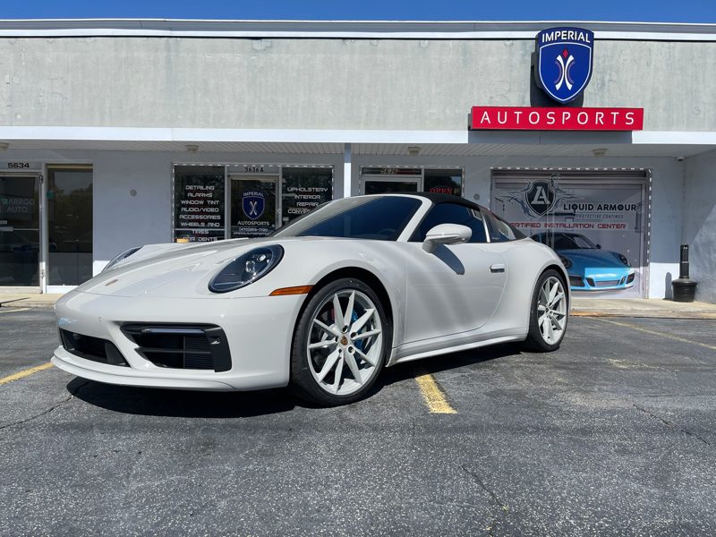 White Porsche 911 Cabriolet parked in front of Imperial Autosports dealership, with a blue car visible inside the showroom.