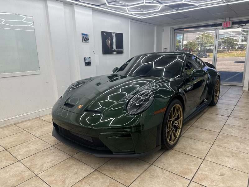 Green Porsche sports car on display in a bright showroom with tiled floor and glass doors in the background. Note: gold wheels and sleek, aerodynamic body.