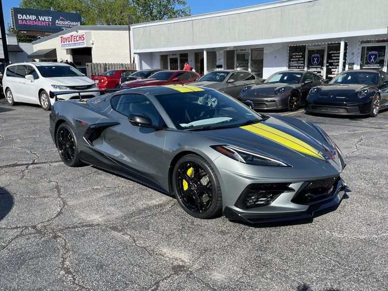 Gray Chevrolet Corvette C8 with a yellow racing stripe, parked in a dealership lot with other cars and storefronts in the background.