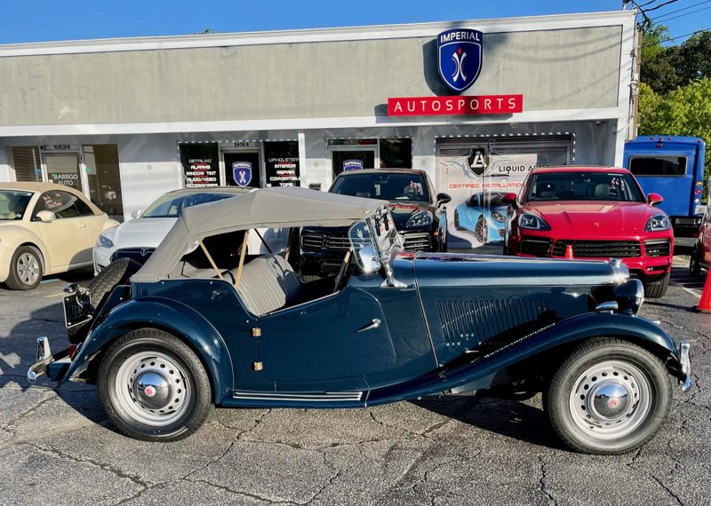 Vintage dark-blue convertible car with a tan soft top parked in front of Imperial Autosports storefront; several modern cars surround it.