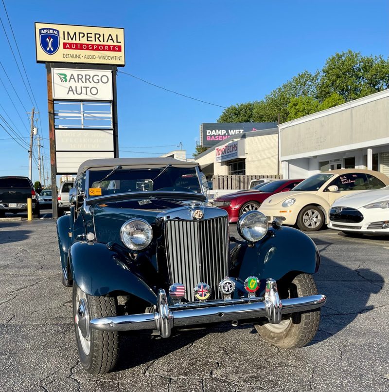 Black 1930s-style vintage convertible with chrome bumper and round headlights, parked in a lot near an Imperial Autosports sign.