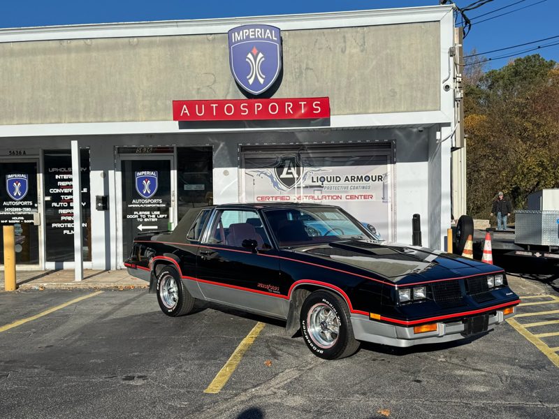 Black late-1980s Pontiac Firebird Trans Am with red pinstripe parked in front of Imperial Autosports storefront. The shop sign and Liquid Armour banner are visible above the entrance.