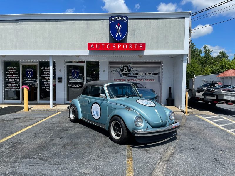 Blue vintage Volkswagen Beetle with a white circular logo on its door parked outside Imperial Autosports on a sunny day