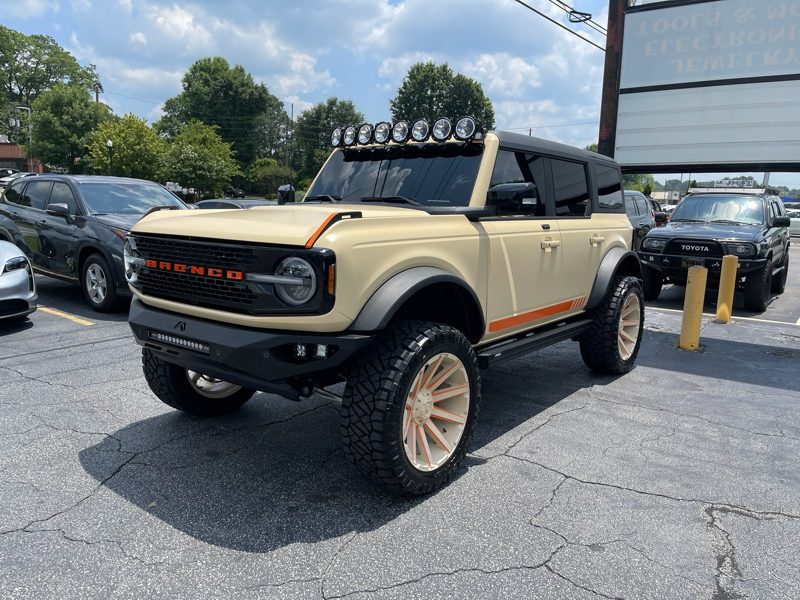 Beige lifted Ford Bronco with orange trim, roof-mounted lights, and large off-road tires in a parking lot at a dealership.