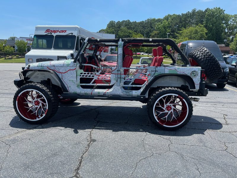 Open-top custom Jeep Wrangler with red interior and map-themed wrap, large red rims, spare tire mounted rear in a parking lot.