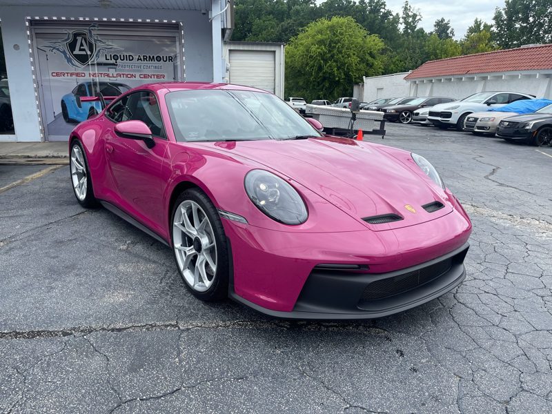 Pink Porsche 911 parked outside a shop with a Liquid Armour banner, featuring silver wheels and a black front bumper.