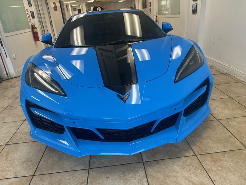 Blue Chevrolet Corvette shown from the front in a showroom, with a black racing stripe down the center and glossy finish.