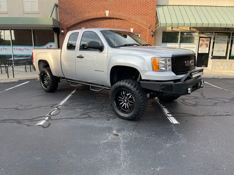 Lifted silver GMC Sierra pickup with oversized off-road tires, black wheels, and running boards parked in a storefront parking lot.