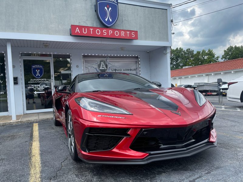 Red Chevrolet Corvette Stingray parked in front of an autosports shop storefront on a sunny day, with the dealership sign visible above.