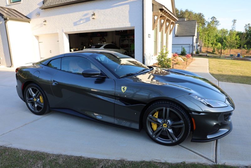 Dark gray Ferrari parked in a suburban driveway in front of a white garage, yellow brake calipers visible through the wheels.