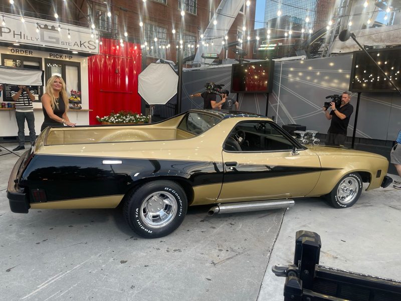 Gold and black classic muscle car on display at a media event, with photographers filming and a woman nearby under string lights and a Philly Steaks sign in the background