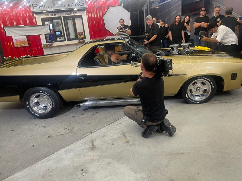 Cinematographer kneels beside a gold-and-black vintage muscle car, filming on a studio set with crew, lights, and onlookers in the background.