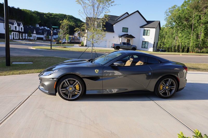 Gray Ferrari sports car parked in a suburban driveway with modern white houses in the background.