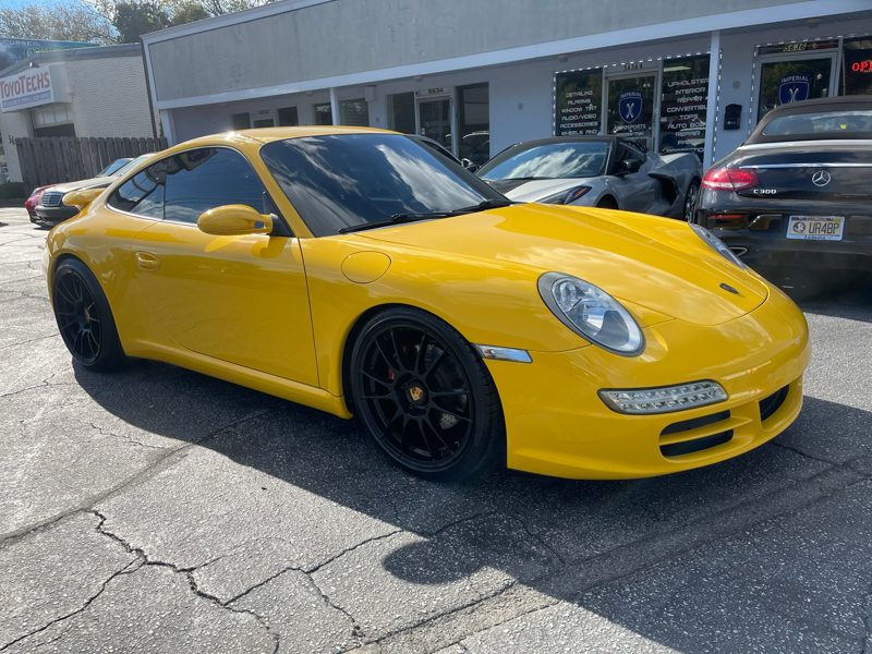 Front-side view of a bright yellow Porsche 911 with black wheels parked in a dealership lot on a sunny day.