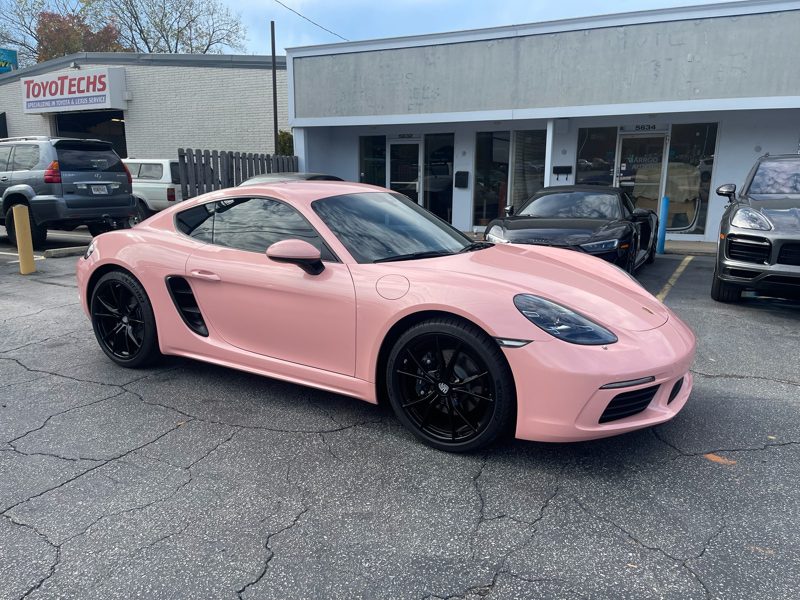Pink Porsche Cayman sports car parked in a car lot beside a storefront with other vehicles nearby.