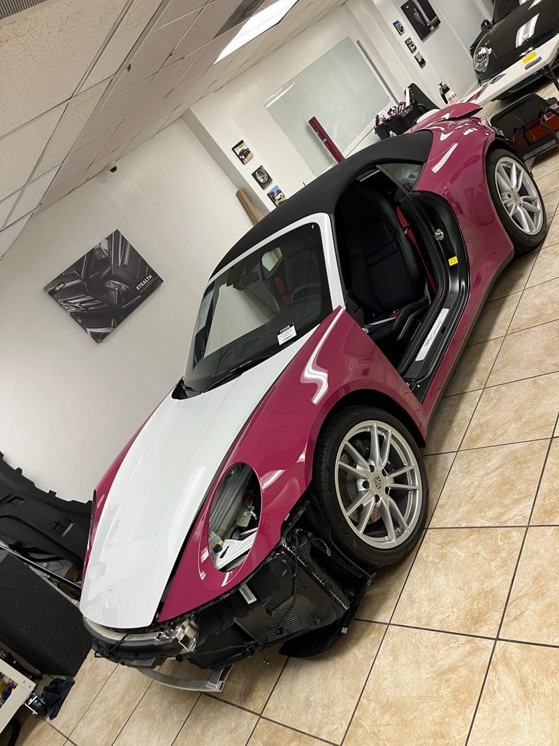 Pink and white sports car with door open in a showroom; front bumper removed, exposing engine area and radiator nearby in a workshop-like space.