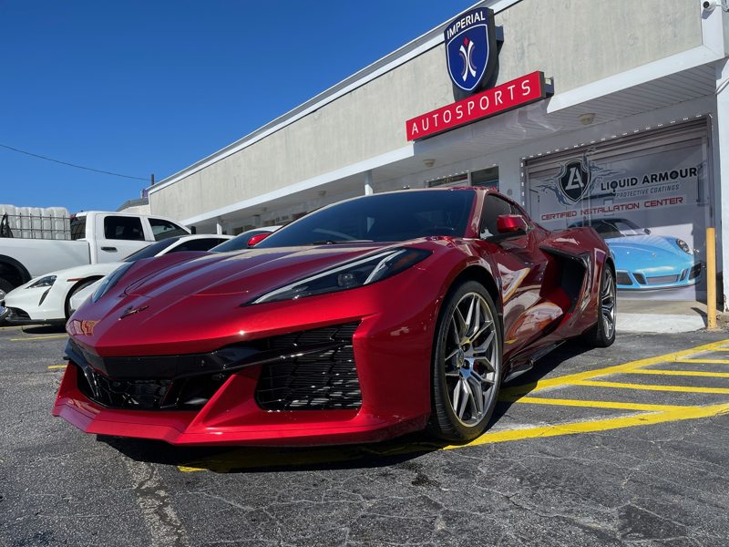Red Chevrolet Corvette C8 parked diagonally in front of Imperial Autosports showroom sign on a sunny day.