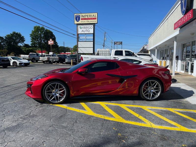 Red two-door sports car with silver wheels parked in a lot in front of an auto shop; yellow diagonal lines on the asphalt and a tall sign in the background.