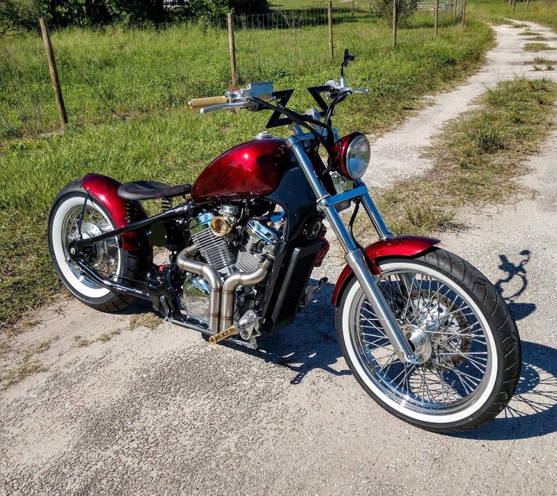 Custom red motorcycle with chrome engine and white-wall tires parked on a rural road, grassy field beside it.