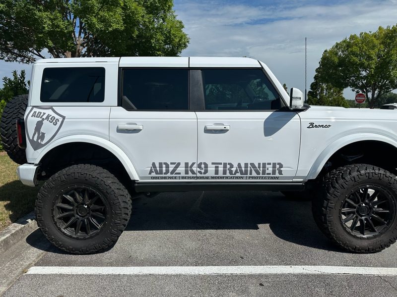 White Ford Bronco SUV with 'ADZ K9 TRAINER' branding parked in a lot; dog-training company vehicle with large off-road tires and badge on the rear quarter panel.