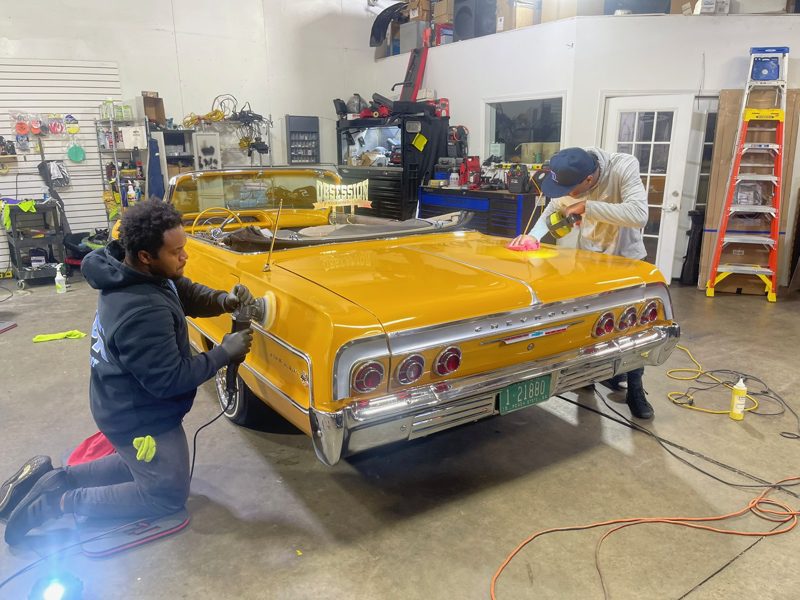 Two workers polish a bright yellow vintage Chevrolet Impala convertible inside a cluttered workshop, using power buffers on the sides and hood.
