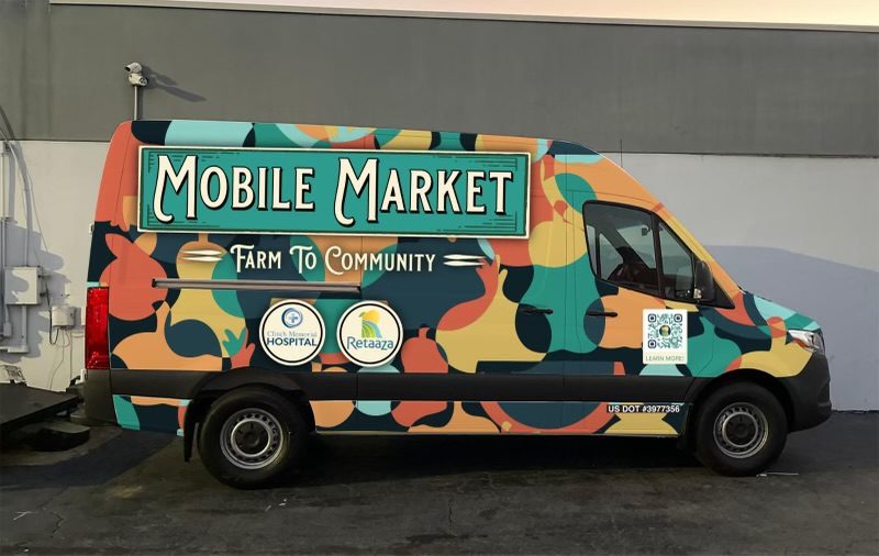 Colorful delivery van with a big 'Mobile Market' sign and 'Farm To Community' slogan, parked beside a gray building.
