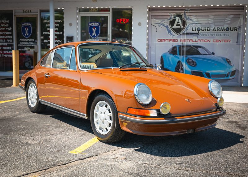 Orange vintage Porsche 911 in a parking lot in front of a storefront with signage including 'Liquid Armour' in the background.