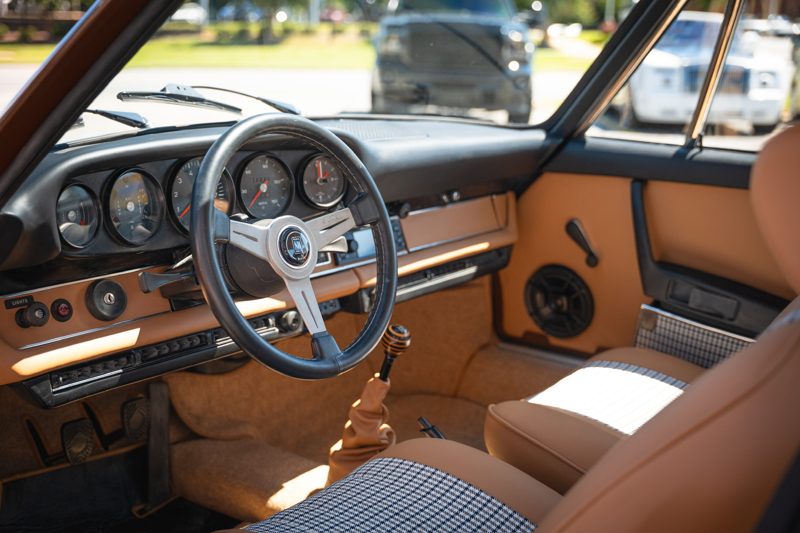 Interior of a classic car: tan dashboard, black steering wheel, and analog gauges with a manual gear shifter in the foreground
