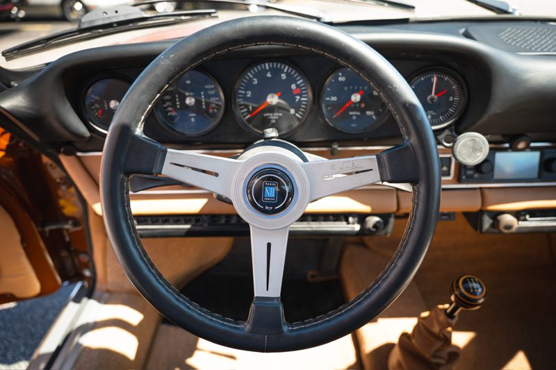 Classic car interior: close-up of a black steering wheel with a silver three-spoke hub, gauges visible in the dashboard, and a brown leather interior in the background.